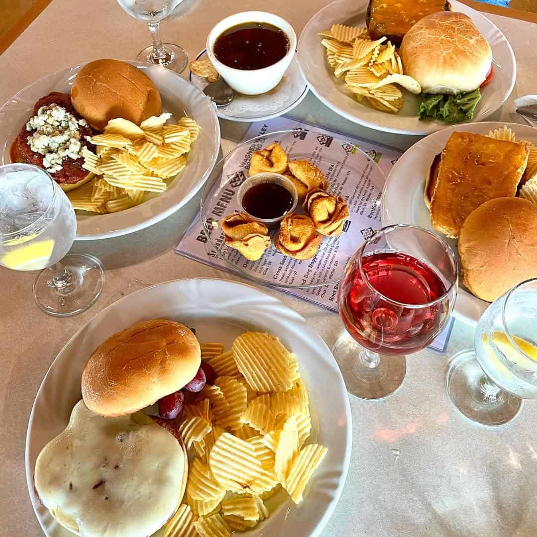 Plates with sandwiches, potato chips, grapes, sauces, pastries, and glasses of water and red drink on a table evoke the inviting spreads often found at Finger Lakes wineries with restaurants.