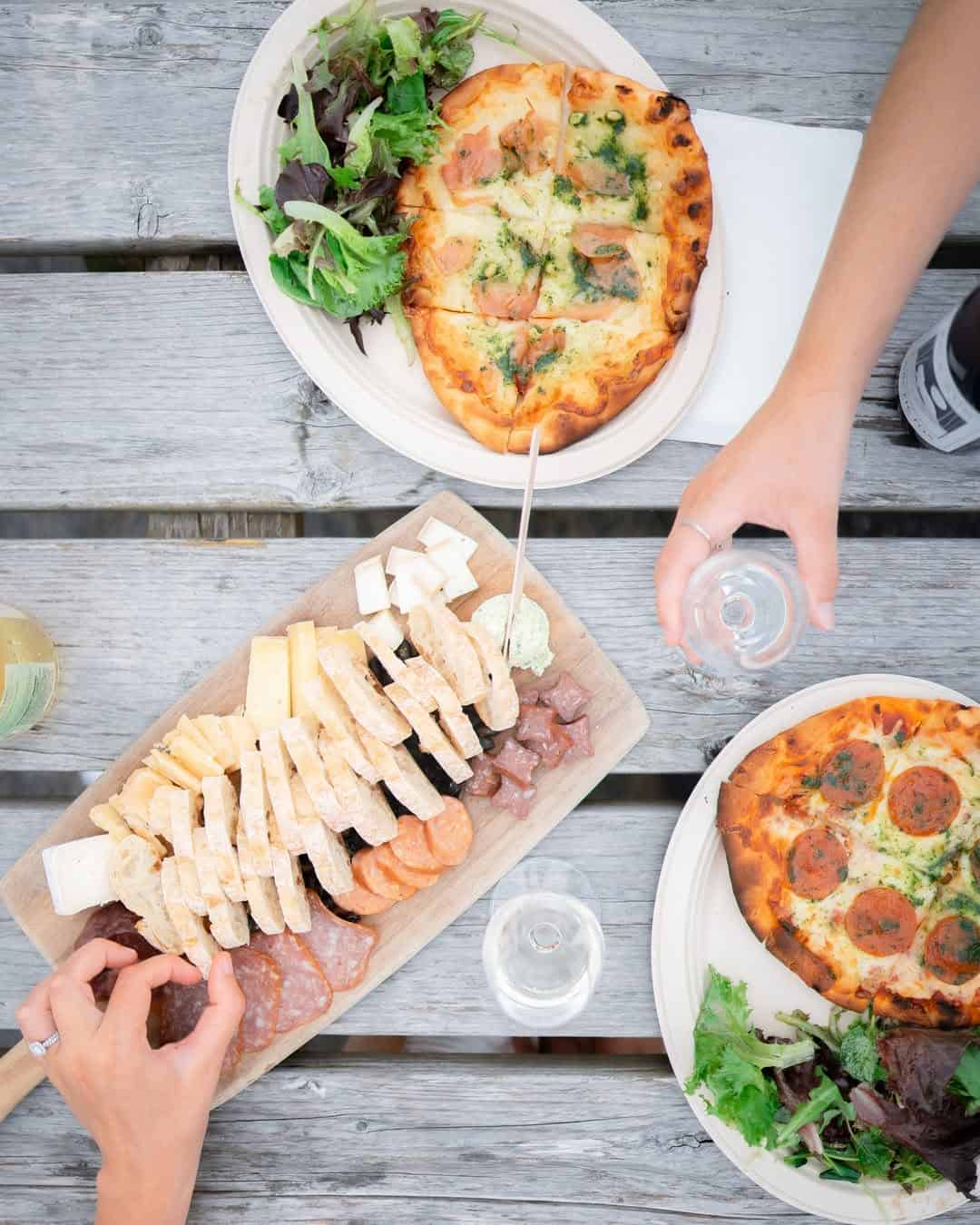 Two people share flatbreads, salad, and a charcuterie board with bread, cheese, and meats on a rustic table&mdash;an experience reminiscent of dining at Finger Lakes wineries with restaurants.