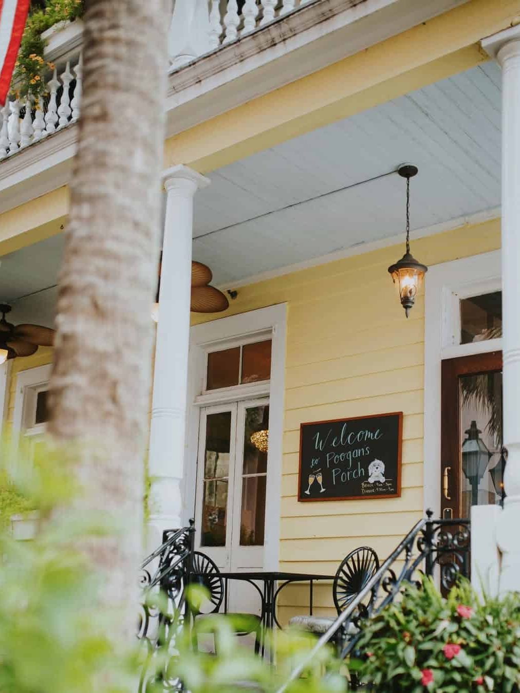 A yellow porch with white columns, a chalkboard sign reading “Welcome to Poogan’s Porch,” and tables and chairs—a charming spot to include on your list of things to do in Charleston with kids.