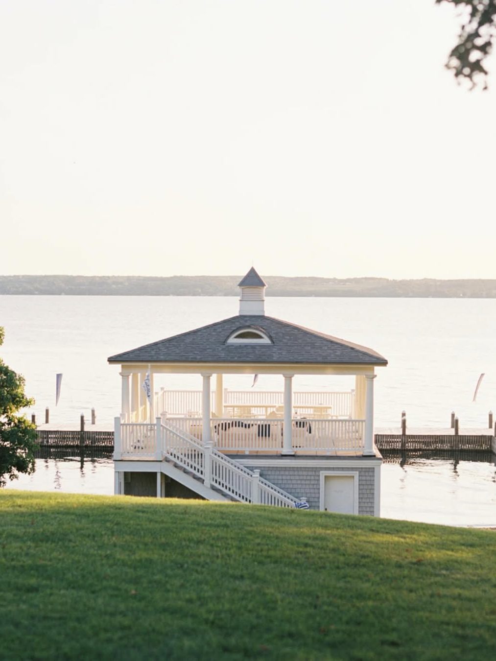 A white gazebo on a pier over calm water, viewed from a grassy hill under soft sunlight—one of the best places to stay in Finger Lakes near wineries.