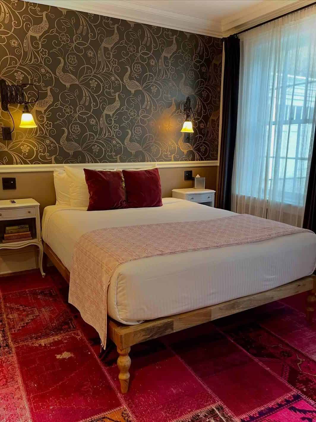 Cozy bedroom with a double bed, red pillows, pink throw, patterned wallpaper, and two wall lamps—one of the best places to stay in Finger Lakes near wineries for a relaxing getaway.