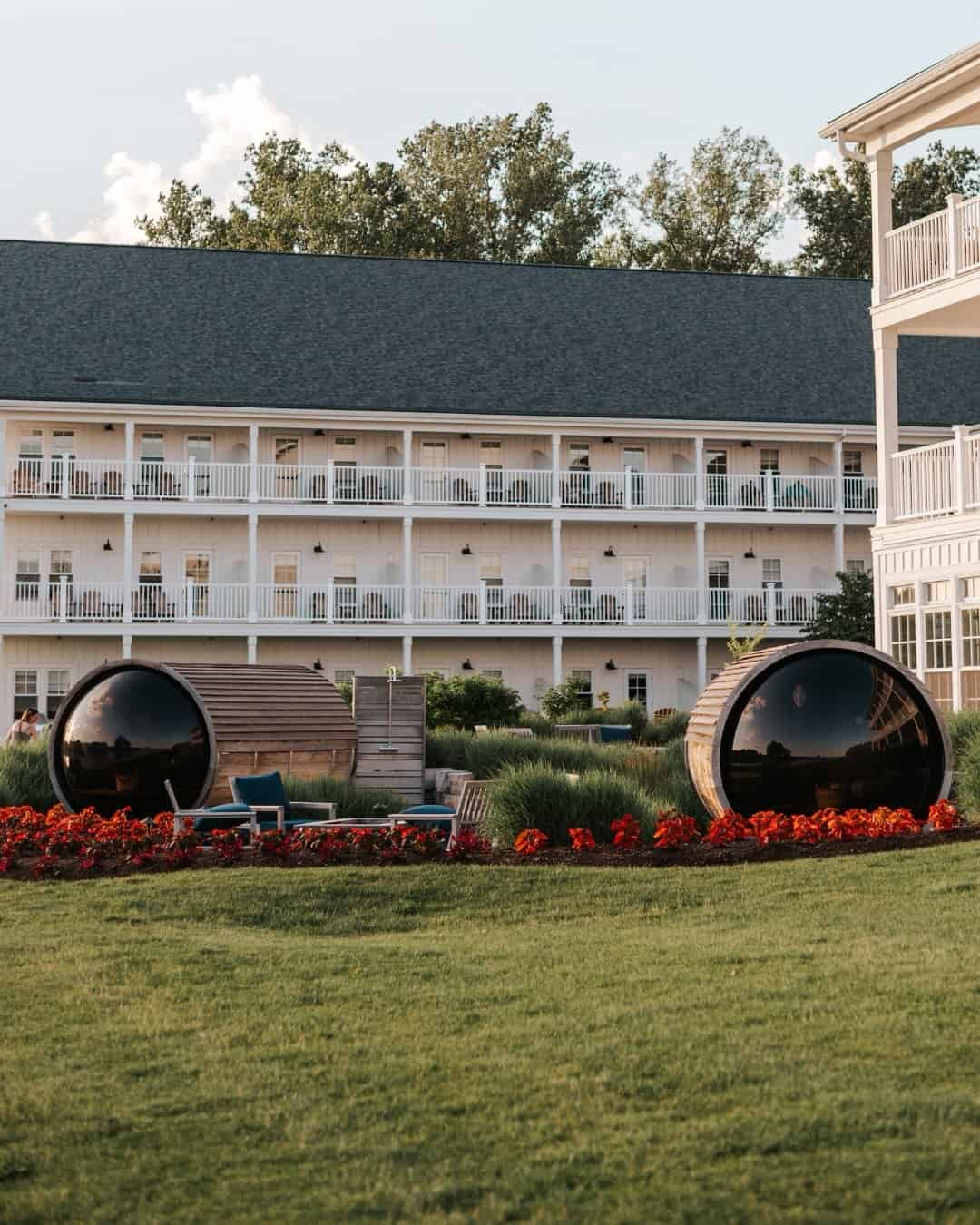 Large white building with balconies, two barrel-shaped saunas, red flowers, and a green lawn in front—one of the Best Places to Stay in Finger Lakes Near Wineries for a relaxing getaway.