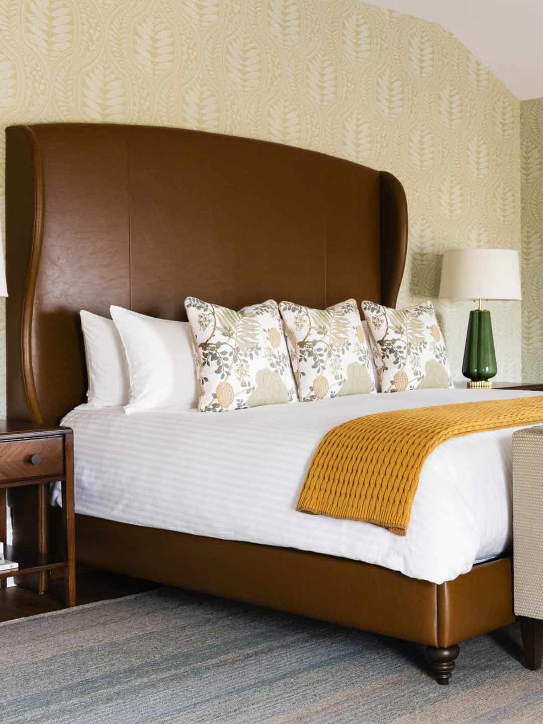 Brown upholstered bed with white bedding, patterned pillows, a yellow throw, and side tables with lamps—perfect for those seeking the Best Places to Stay in Finger Lakes Near Wineries.