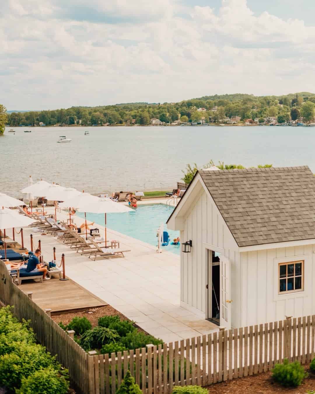 A lakeside pool with lounge chairs, umbrellas, and a small white building beside the water on a sunny day—one of the best places to stay in Finger Lakes near wineries.