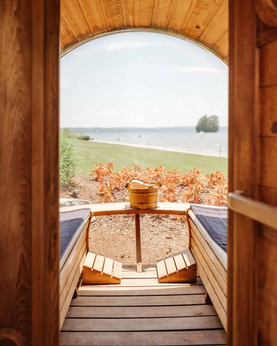 Wooden sauna with benches, looking out to a lake and grassy shore through an arched doorway—one of the Best Places to Stay in Finger Lakes Near Wineries for a serene retreat.