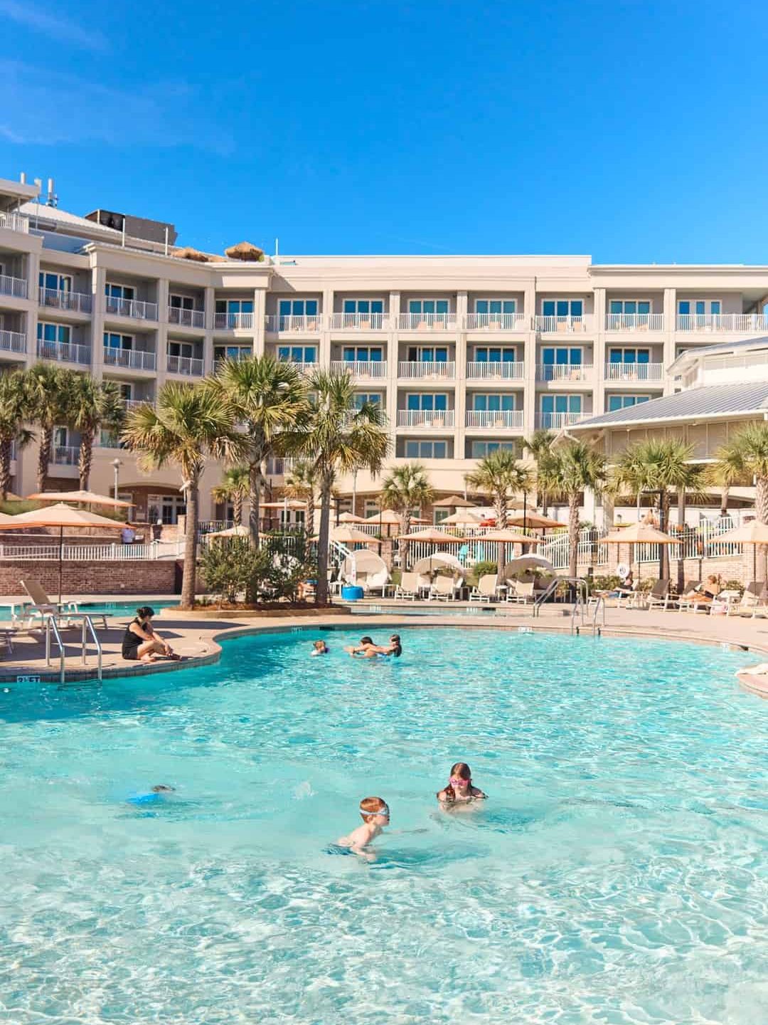 People swim and relax in a resort pool surrounded by palm trees and a hotel building under a clear blue sky, making it one of the best things to do in Charleston with kids.