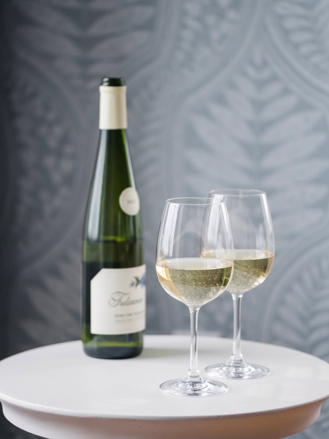 A wine bottle and two glasses filled with white wine on a white table against a patterned gray background, evoking the relaxing ambiance of the Best Places to Stay in Finger Lakes Near Wineries.