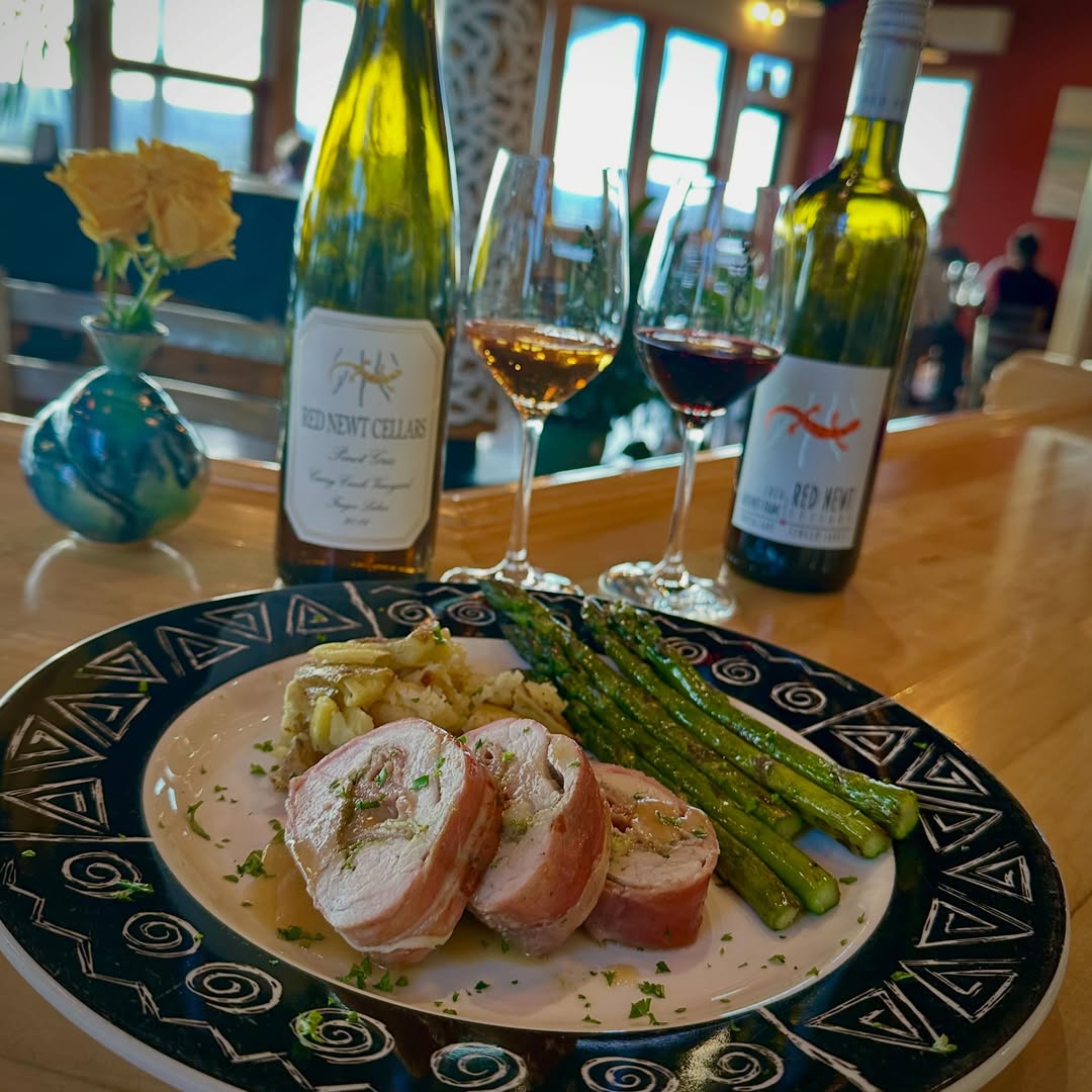 A plated meal with stuffed chicken, asparagus, and potatoes, set against wine bottles and glasses&mdash;perfect for enjoying at one of the renowned Finger Lakes wineries with restaurants.