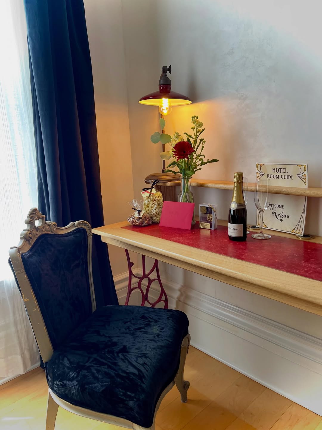 Elegant desk with a black velvet chair, flowers, champagne, and a lamp, set beside a window with dark curtains—perfect for relaxing at one of the Best Places to Stay in Finger Lakes Near Wineries.