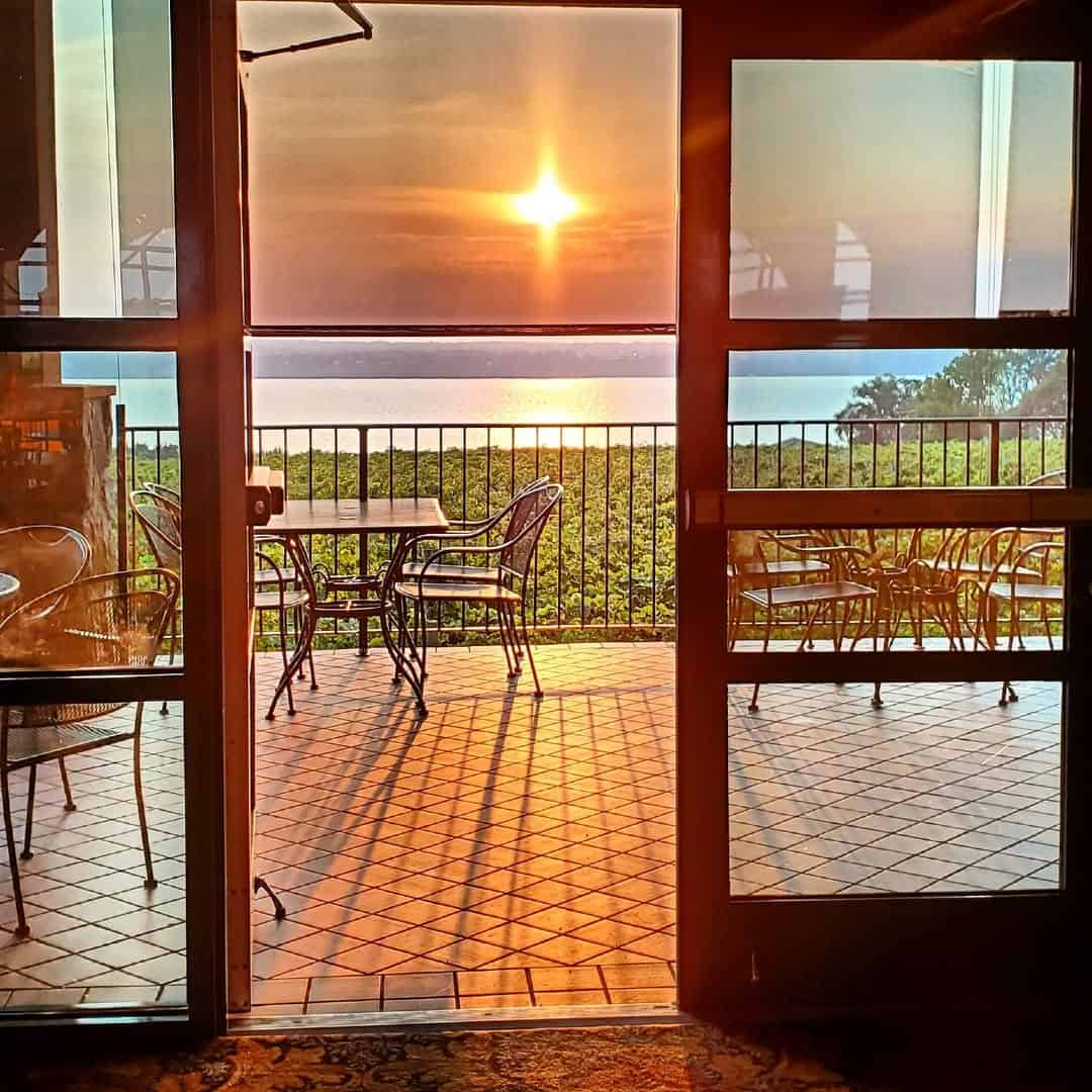 A sunlit patio with empty tables overlooks greenery and water at sunset, viewed through glass doors&mdash;capturing the relaxing atmosphere found at many Finger Lakes wineries with restaurants.