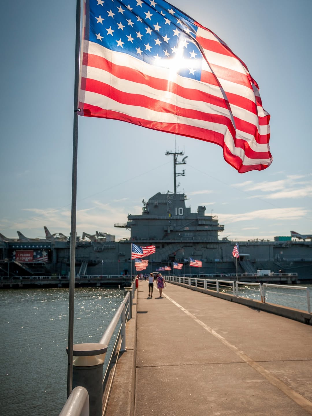 American flag waving in the foreground, with a large navy ship docked at the end of a pier under a sunny sky—one of the top things to do in Charleston with kids for an educational and memorable family outing.