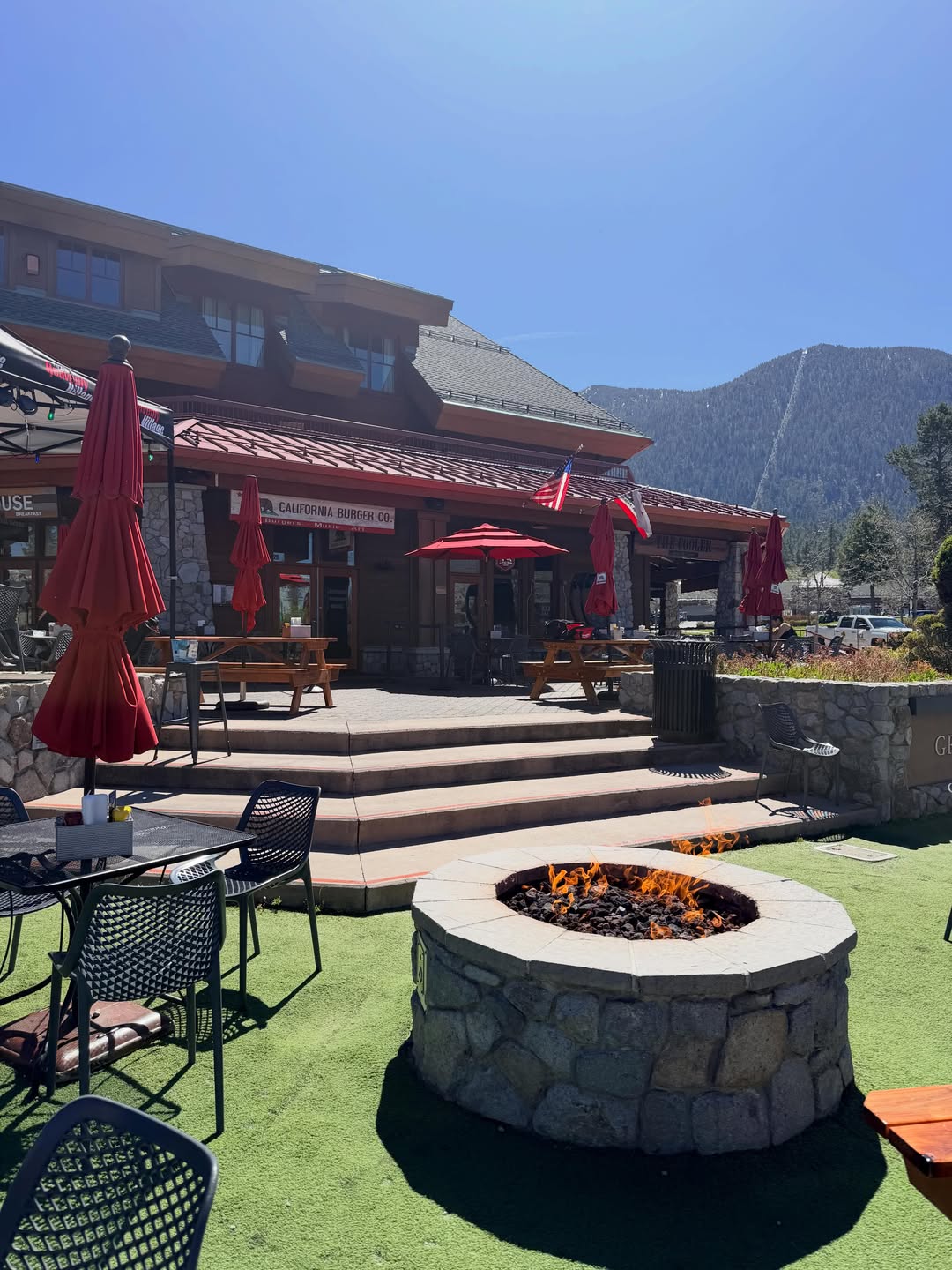 Outdoor seating area with red umbrellas and a fire pit in front of a building, with mountains in the background—perfect for relaxing during your family vacation to Lake Tahoe.