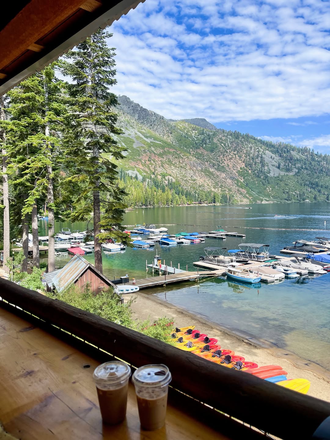 Two iced coffees sit on a wooden table, overlooking boats docked at a lake with mountains and pine trees in the background—a perfect moment from a family vacation to Lake Tahoe.