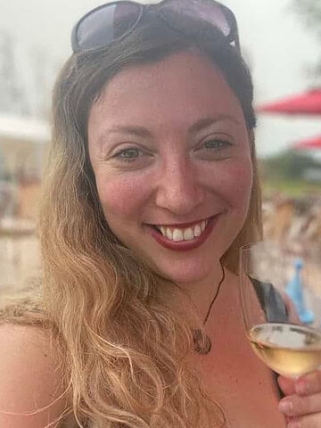 Smiling woman with wavy hair holding a glass of white wine outdoors, sunglasses on her head, enjoying one of the Finger Lakes Wine Tours.