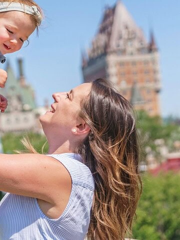 A smiling woman lifts a happy toddler in front of a historic castle-like building on a sunny day.