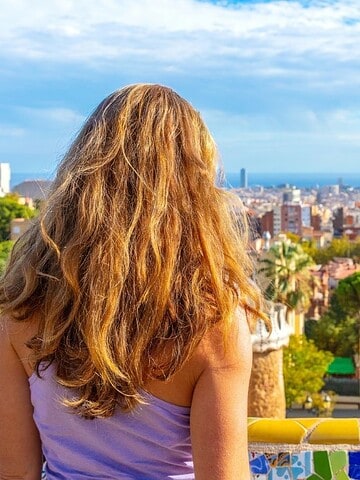 Woman with long hair looks out over Barcelona from a mosaic balcony in Park Güell on a sunny day.