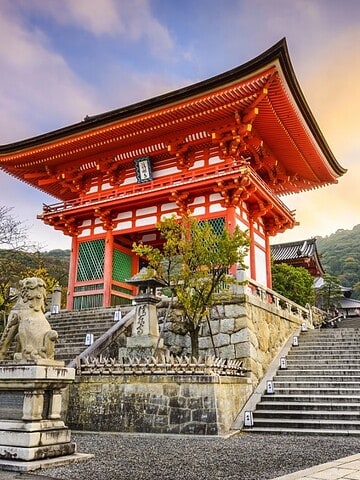 Red pagoda entrance and stone steps at Kiyomizu-dera Temple in Kyoto, Japan, with a sunset sky in the background.
