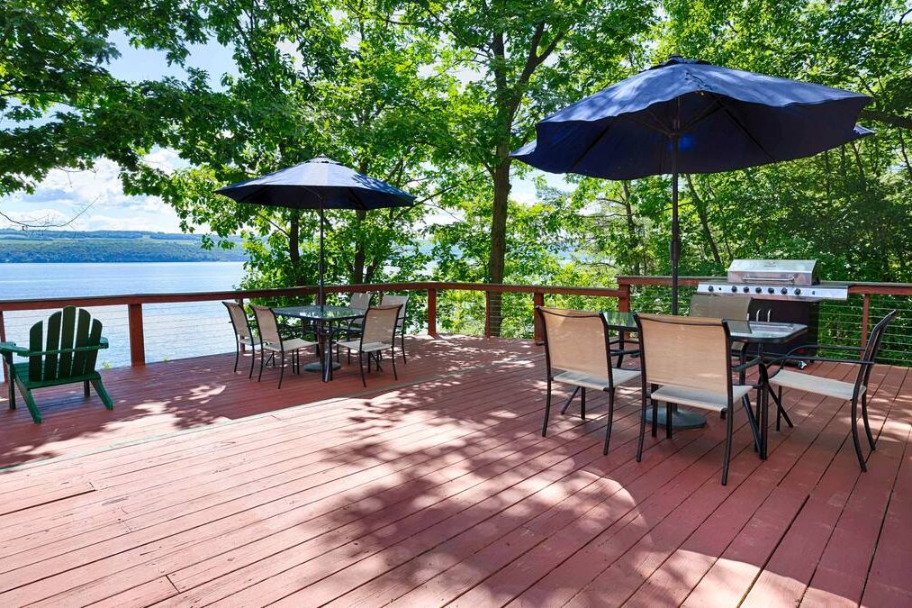 Spacious wooden deck at our Finger Lakes Vacation Rentals features tables, chairs, umbrellas, a grill, and stunning lake views surrounded by trees—perfect for relaxing after a day exploring nearby cottages and cabins.