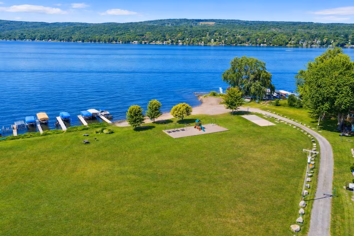 A lakeside park with green grass, a playground, docks, and trees, bordered by a blue lake and distant hills—perfect for relaxing or enjoying nearby Finger Lakes vacation rentals.