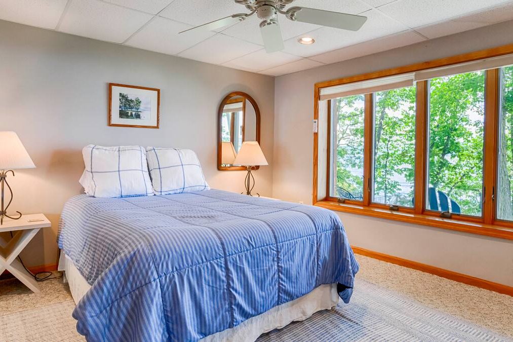 Cozy bedroom with blue bedding, large windows overlooking trees, and soft lighting from two lamps—perfect for relaxing getaways in Finger Lakes Vacation Rentals.