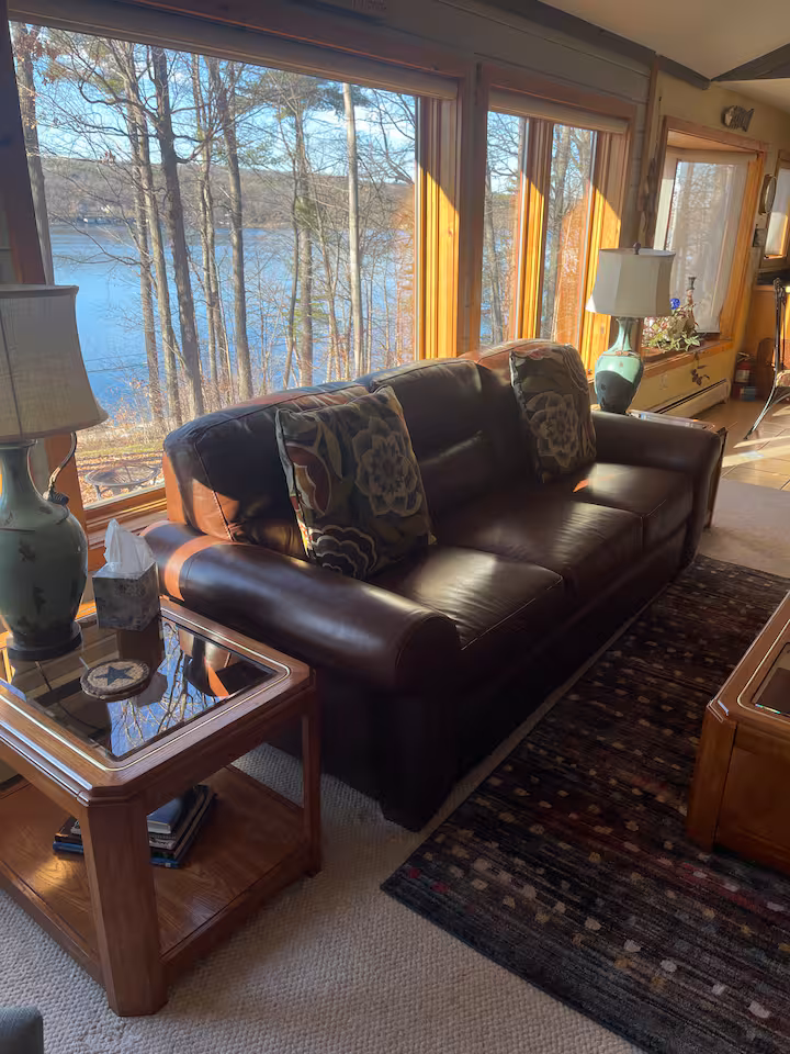 Brown leather sofa with patterned pillows in a sunlit living room, large windows reveal a lake and trees outside—ideal for cozy Finger Lakes vacation rentals, cottages, or cabins.