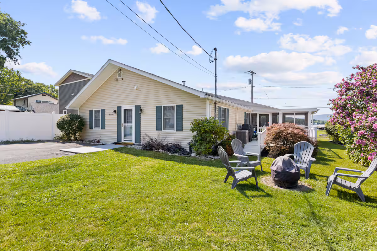 Single-story house with a grassy yard, patio chairs, fire pit, shrubs, and blooming bush under a blue sky—perfect for relaxing Finger Lakes vacation rentals.