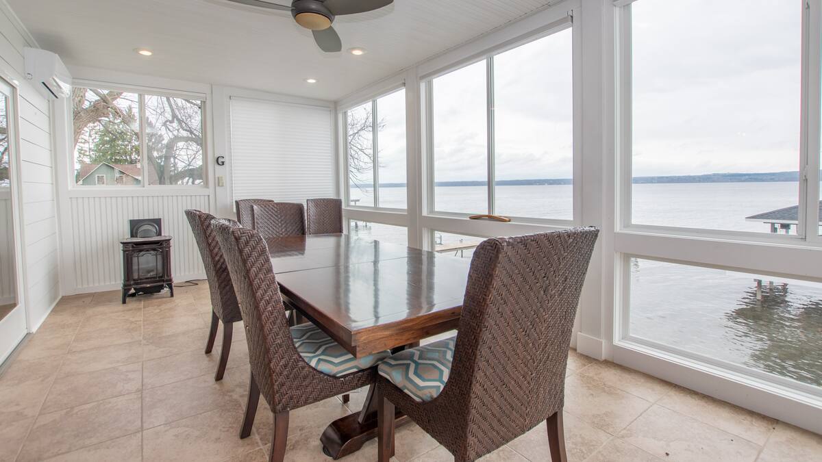 Dining table with six chairs in a bright sunroom overlooks a large lake through floor-to-ceiling windows, perfect for relaxing Finger Lakes vacation rentals.