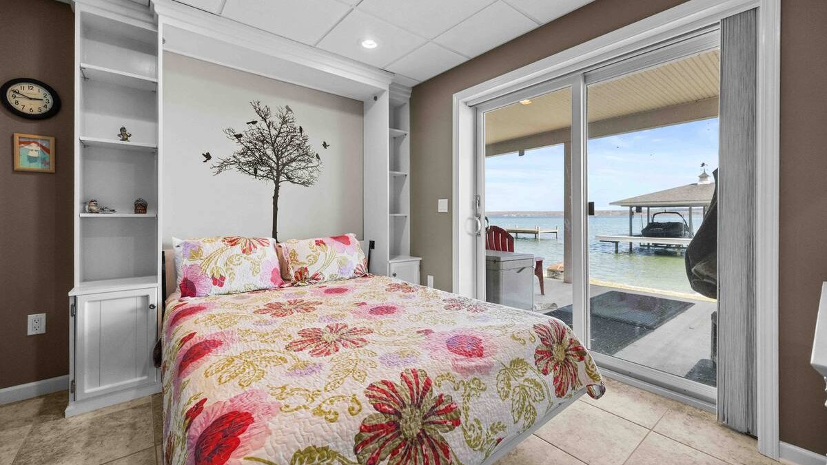 Cozy bedroom with floral bedding, a tree decal, and sliding glass doors overlooking a lakeside dock—perfect for relaxing Finger Lakes vacation rentals.