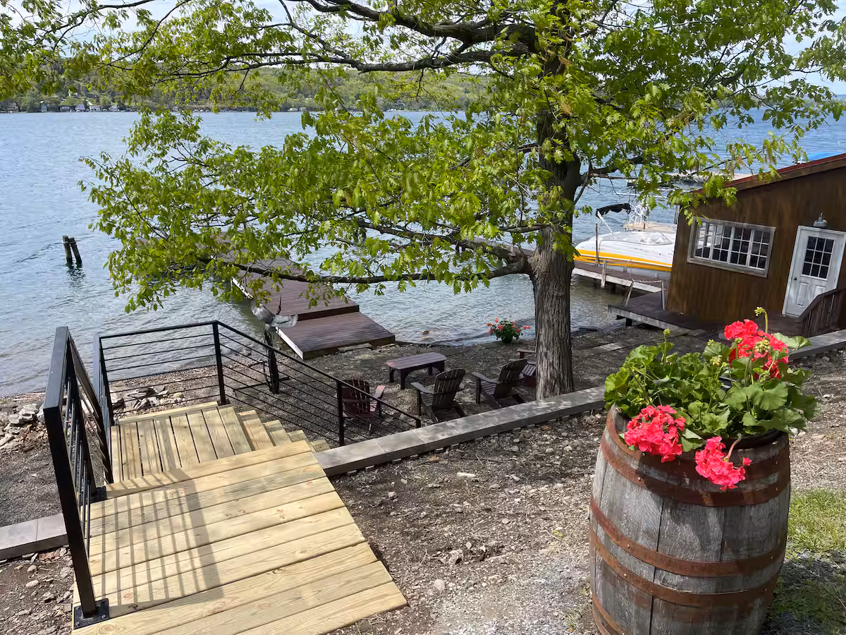 A lakeside scene with cozy cottages, a dock, Adirondack chairs, a tree, and a barrel planter with pink flowers—perfect for relaxing Finger Lakes vacation rentals.