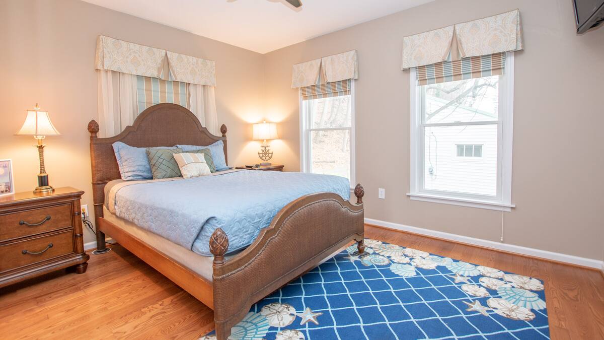 A cozy bedroom reminiscent of charming cabins, featuring a wooden bed, blue bedding, a patterned rug, and two windows with valances.