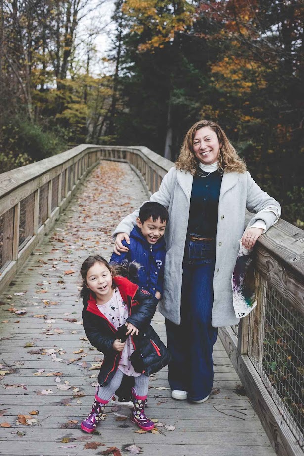 A woman and two children smile on a wooden bridge surrounded by autumn trees, enjoying the beauty near Finger Lakes vacation rentals.