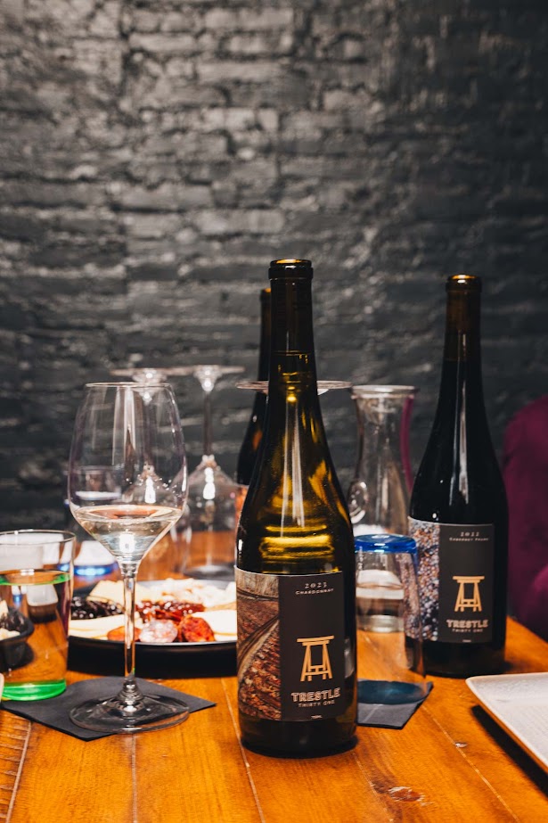 Two wine bottles, glasses, and plates with snacks on a wooden table set against a dark brick wall evoke the cozy charm of Finger Lakes Wine Tours.