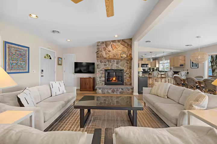 Cozy living room with beige sofas, stone fireplace, coffee table, and open kitchen in the background—perfect for relaxing in one of our inviting Finger Lakes vacation rentals or charming cottages.
