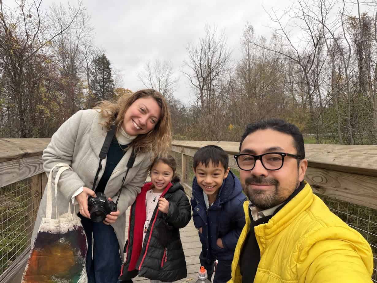 A smiling family of four poses on a wooden bridge outdoors on a cloudy day, with trees in the background—capturing memories while exploring the Best Places to Stay in Finger Lakes Near Wineries.
