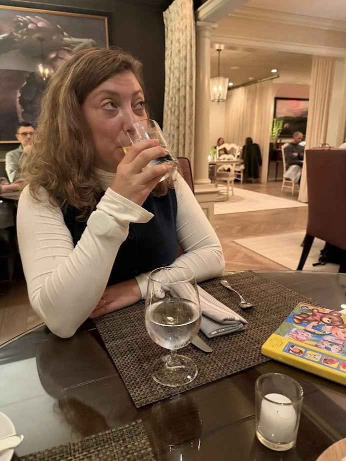 Woman sitting at a restaurant table, enjoying a drink—perhaps inspired by Finger Lakes Wine Tours—with a water glass and a kids’ activity book nearby.