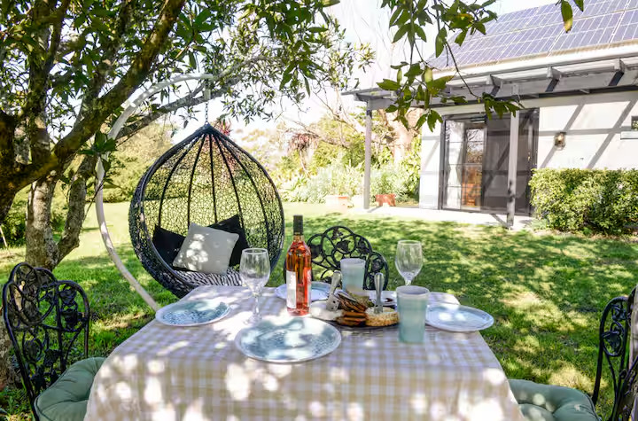 Table set for four with wine and snacks in a sunny garden, near a hanging chair and a house with solar panels—perfect for relaxing at cottages or Finger Lakes vacation rentals.