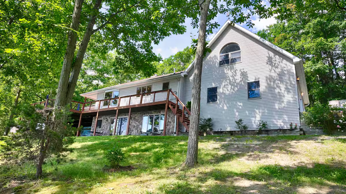 A two-story house with large windows, a wooden deck, and surrounded by trees on a grassy slope—perfect for those seeking cozy cottages or Finger Lakes vacation rentals.