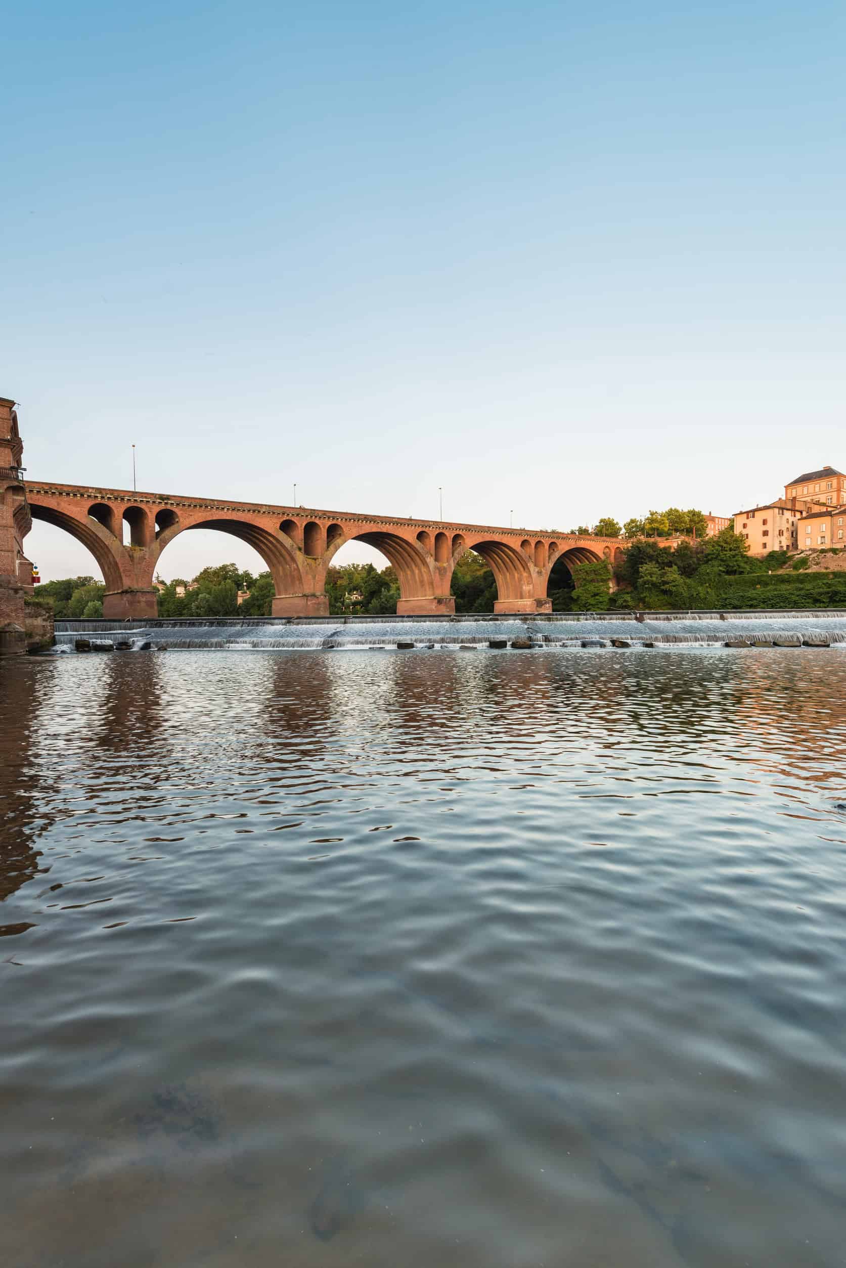 Stone arched bridge over a calm river with reflections, trees, and buildings under a clear sky—perfect scenery for a luxury barge cruise passing gently beneath.