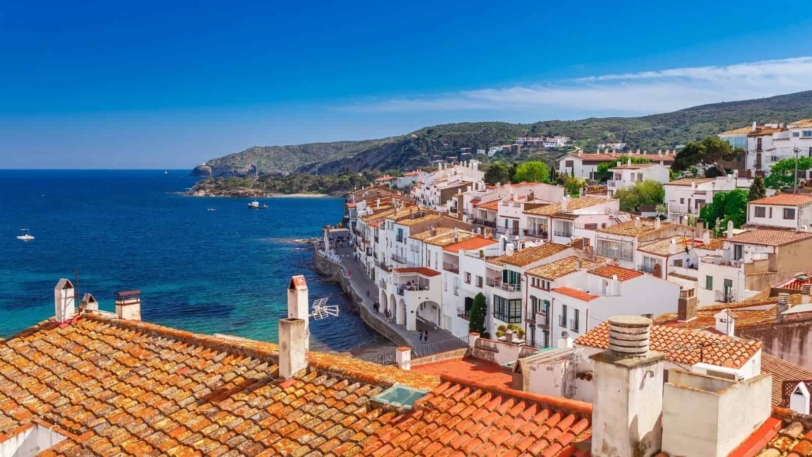 Red-tiled roofs of seaside village with white houses beside blue sea under a clear, sunny sky.