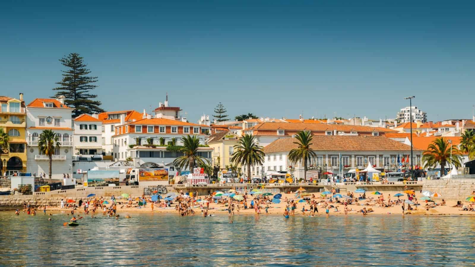 Crowded beach with colorful umbrellas, people swimming, and seaside buildings with red roofs and palm trees.