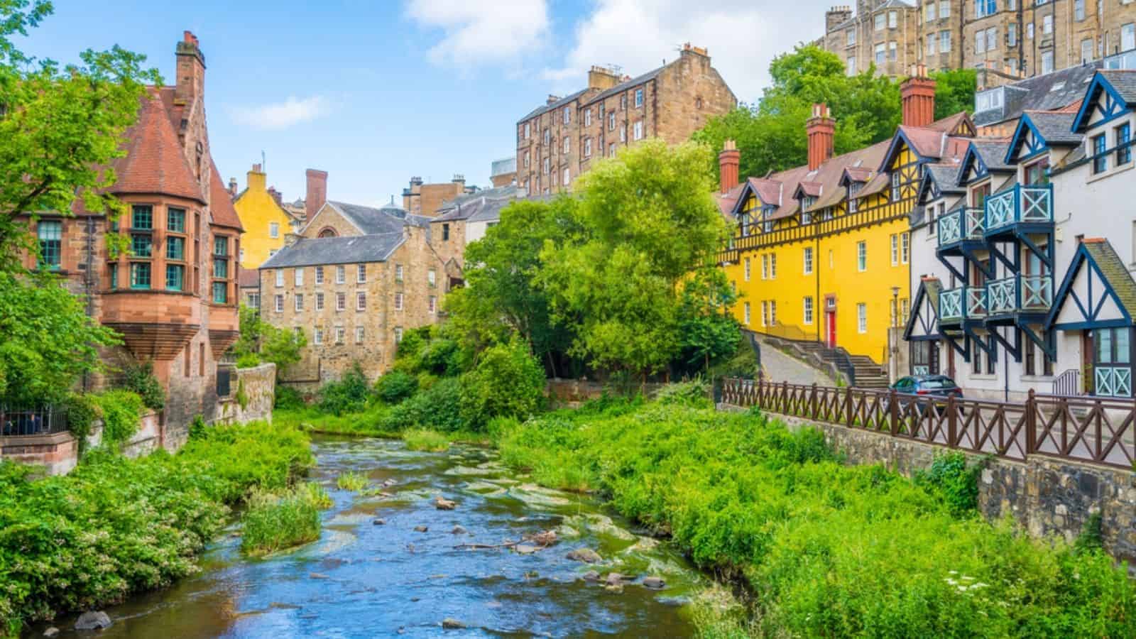 Colorful historic buildings line a lush green riverside under a bright blue sky with scattered clouds.