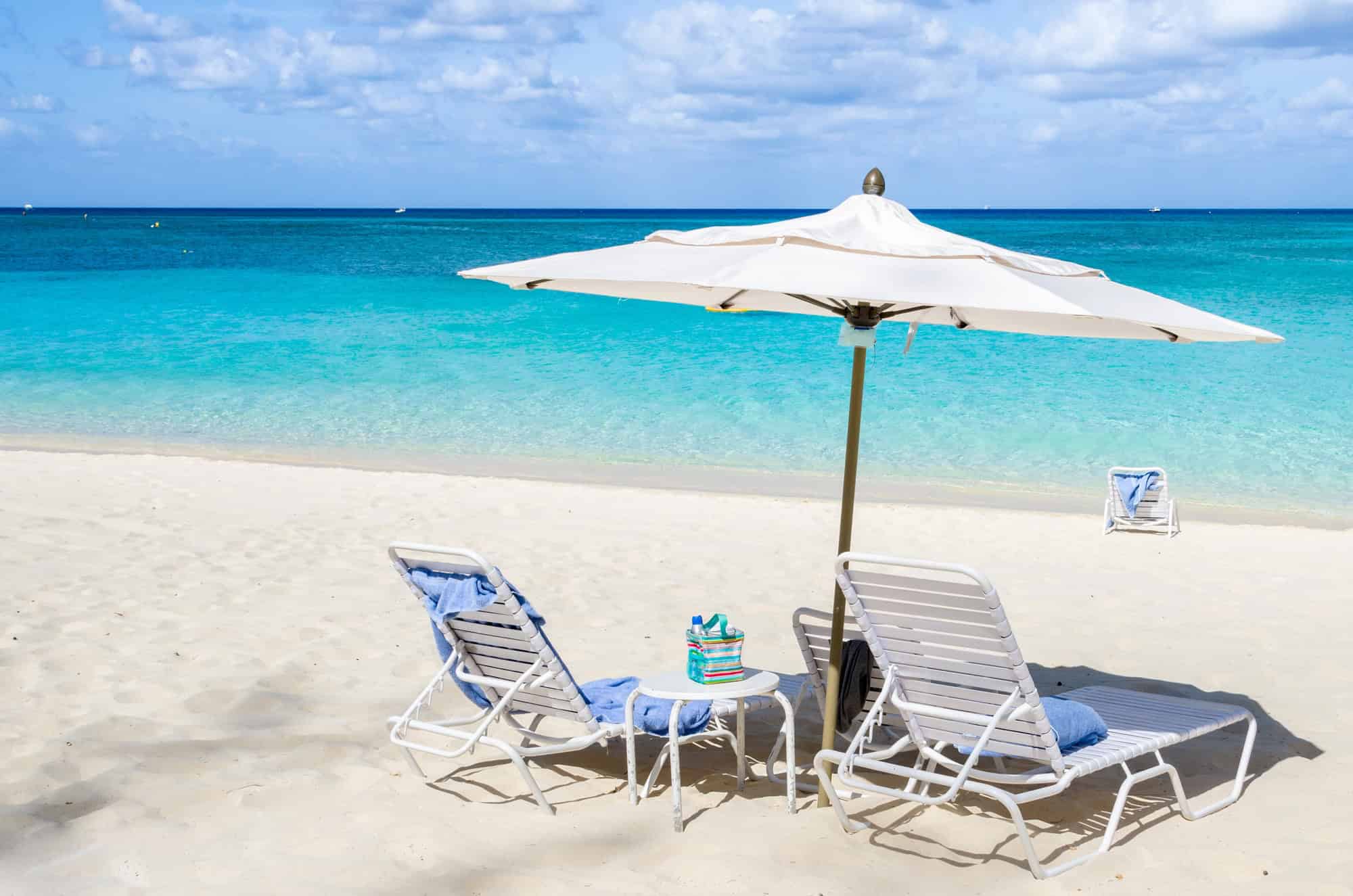 Two lounge chairs and an umbrella on a sandy beach facing clear blue ocean water under a sunny sky, capturing the essence of Grand Cayman luxury travel.
