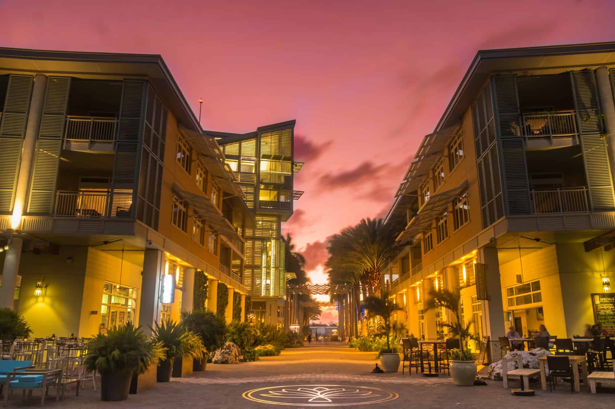 Outdoor dining area between two modern buildings at sunset with a pink sky and palm trees in the distance, evoking the ambiance of Grand Cayman luxury travel.