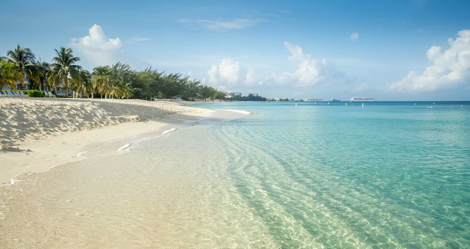 Clear turquoise water and a white sand beach with palm trees under a bright blue sky with scattered clouds create the perfect setting for Grand Cayman luxury travel.