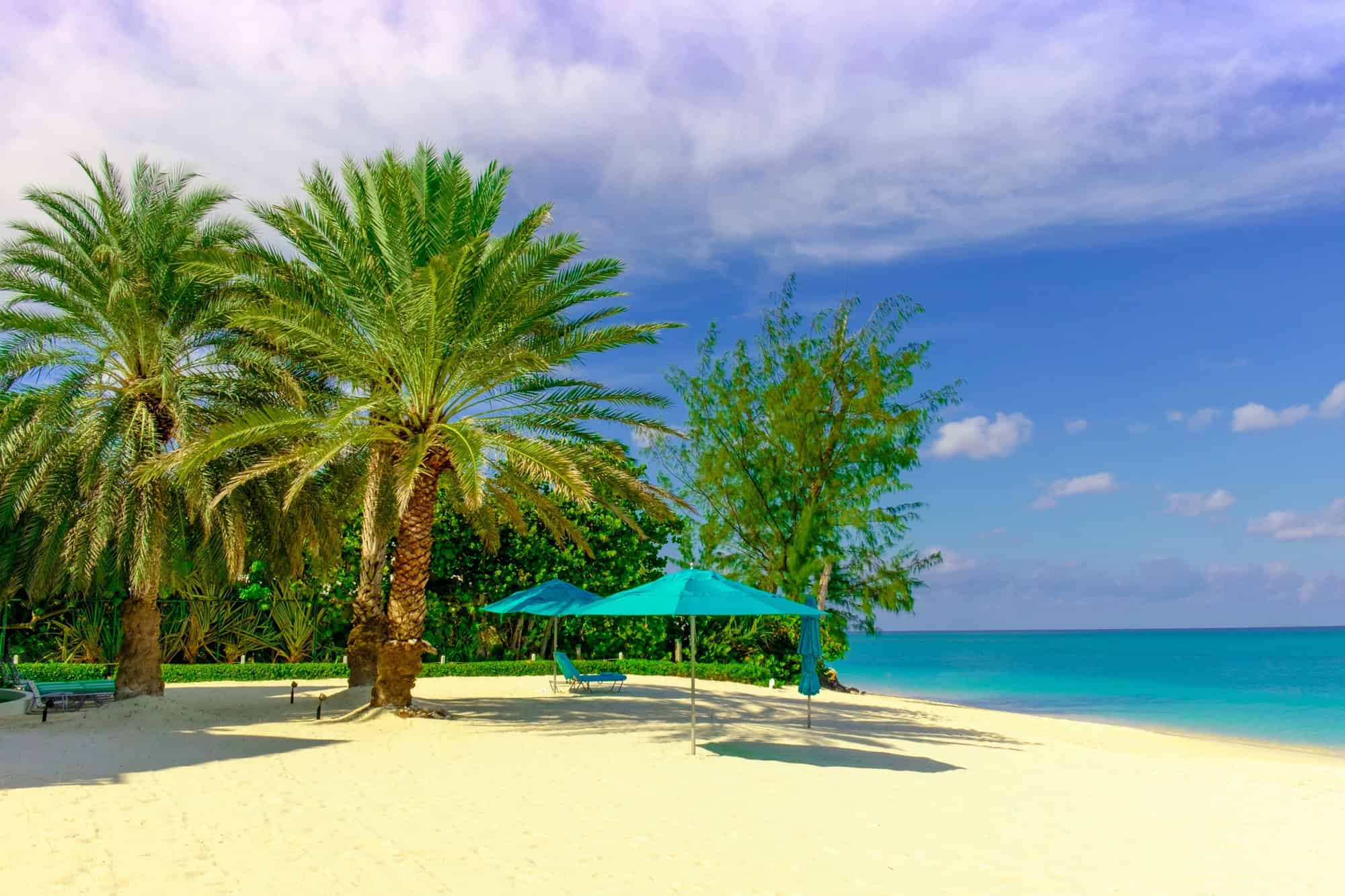 Palm trees and blue umbrellas on a sandy beach beside the clear turquoise sea set the scene for Grand Cayman luxury travel under a sunny, partly cloudy sky.