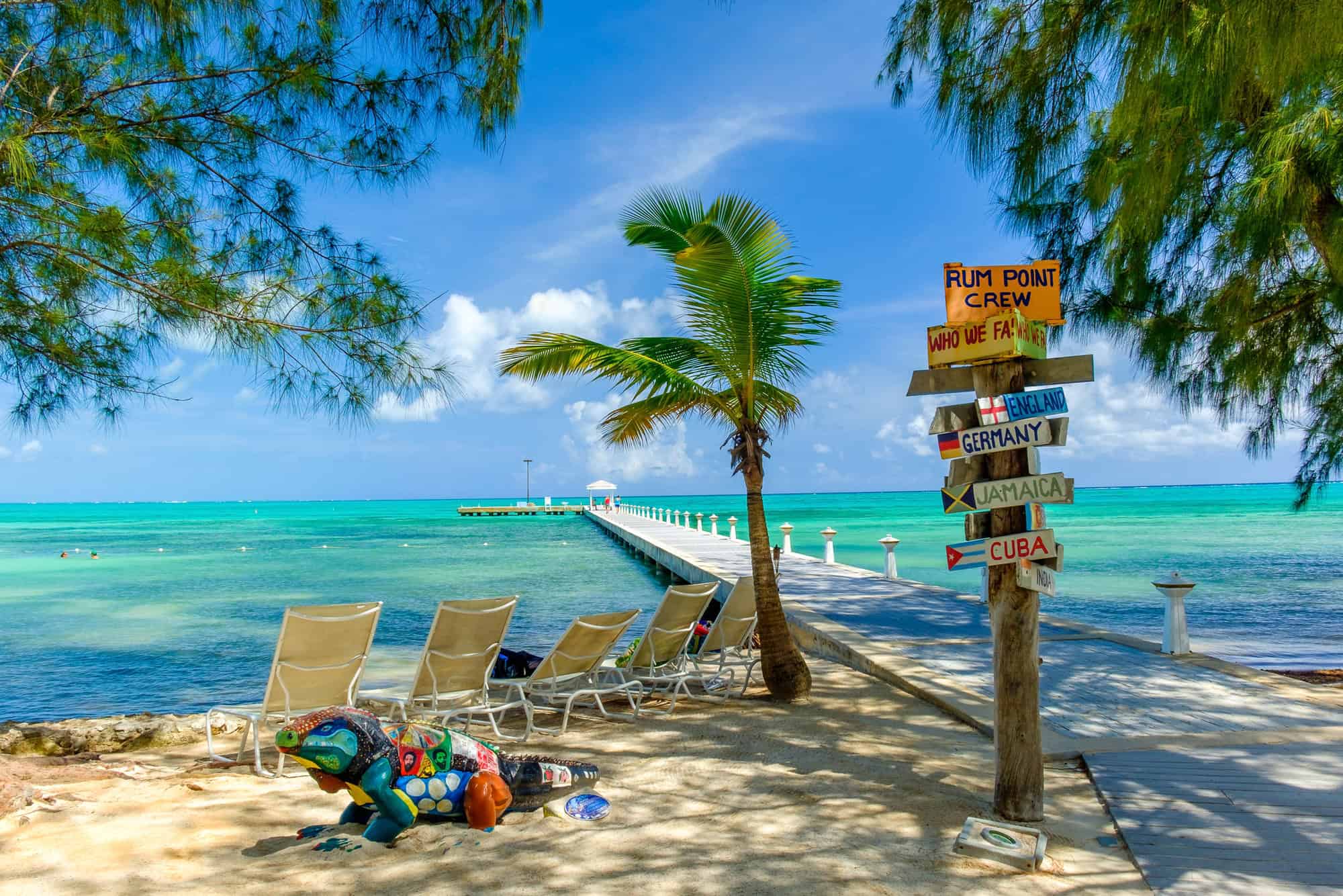 Tropical beach with lounge chairs, a colorful lizard sculpture, signposts, palm, and a long pier over blue water&mdash;perfect for those seeking the ultimate Grand Cayman luxury travel experience.