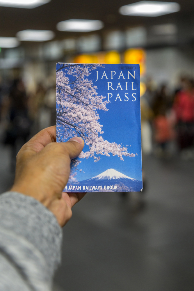 A hand holds a Japan Rail Pass booklet with cherry blossoms and Mount Fuji, capturing the excitement of traveling in Japan inside a busy station.