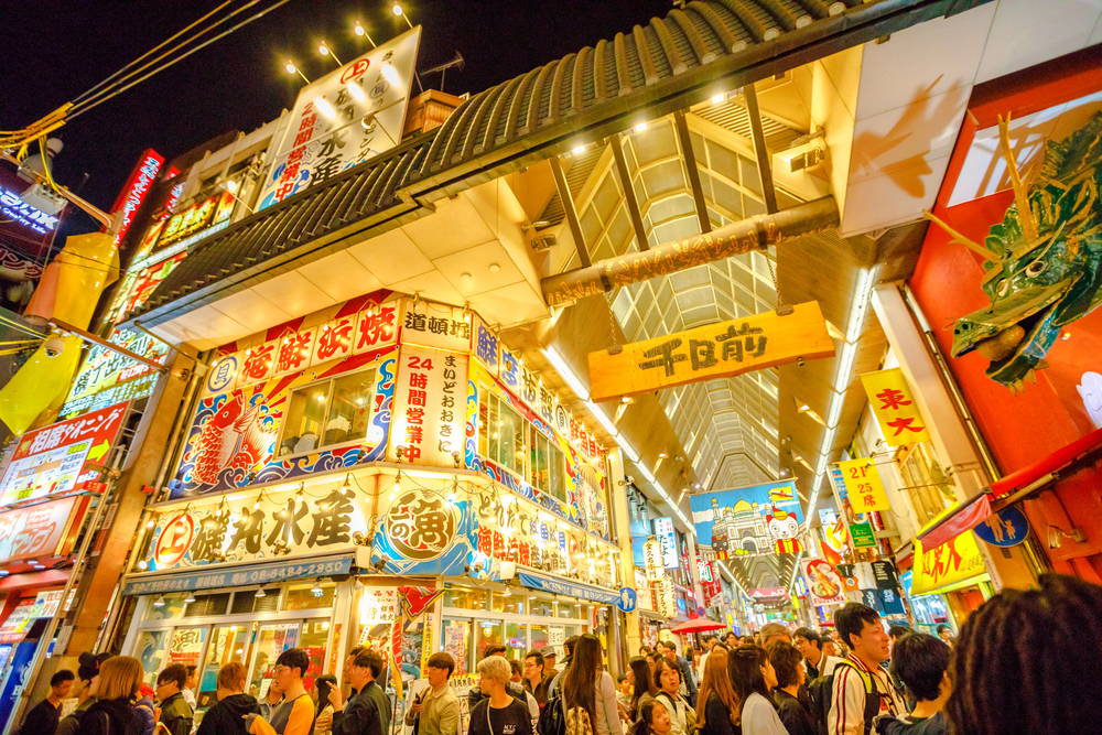 Crowded night market street in Japan with bright signs, shops, and people walking under illuminated lights—a quintessential scene when traveling in Japan.