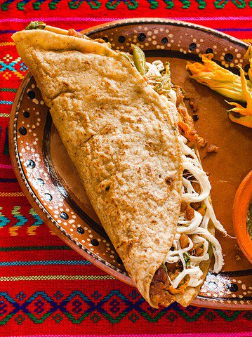 A traditional Mexican quesadilla with cheese, salsa, and squash blossoms sits on a colorful woven tablecloth—a must-try for those seeking authentic Foodie Destinations.