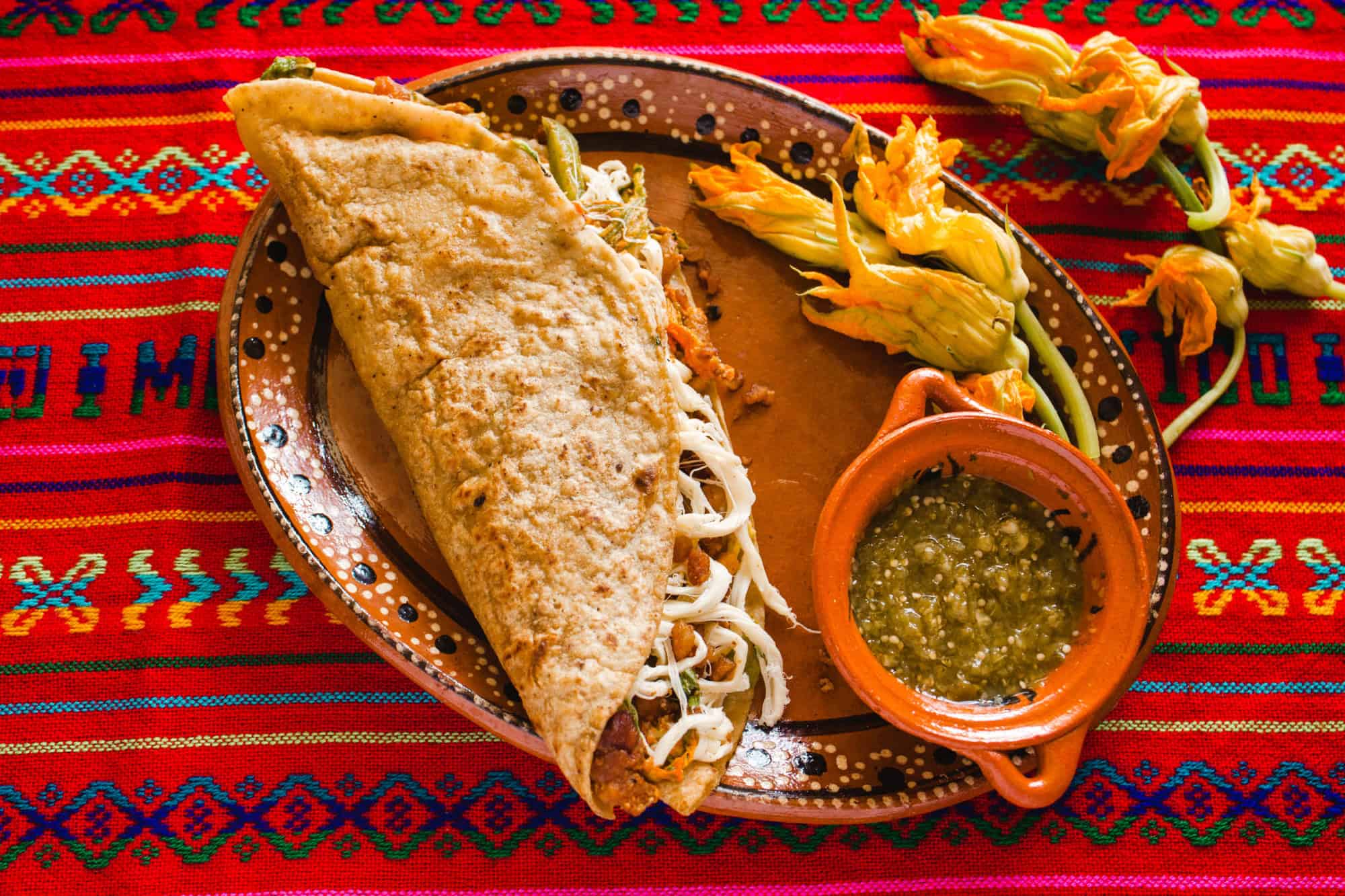 A traditional Mexican quesadilla with cheese, salsa, and squash blossoms sits on a colorful woven tablecloth—a must-try for those seeking authentic Foodie Destinations.